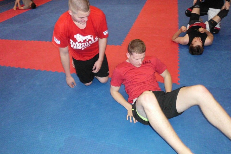 Zwei Sportler in roter Kleidung &uuml;ben auf einer blauen Matte, w&auml;hrend im Hintergrund weitere Personen trainieren.