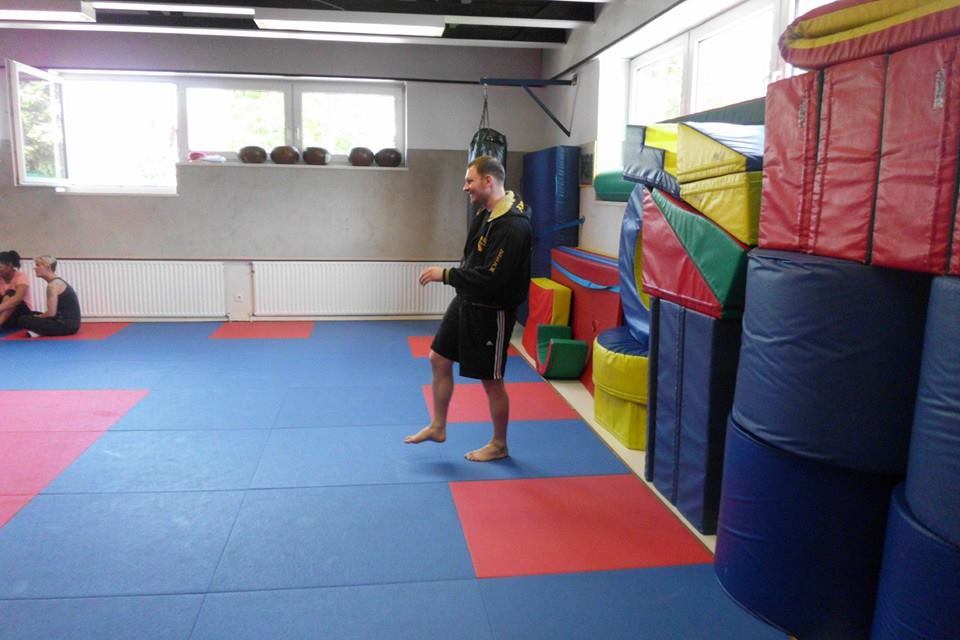 Mann in Sportkleidung steht auf einer bunten Matte in einem Trainingsraum mit gepolsterten Ger&auml;ten und Fenster.