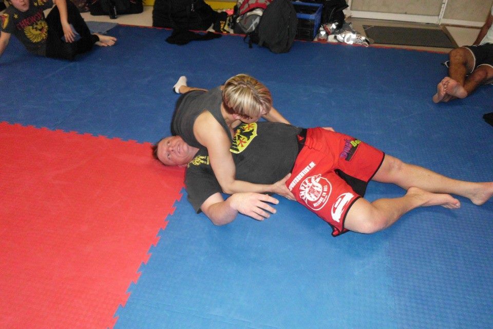 Zwei Personen im Kampfsporttraining; eine Person sitzt auf einer liegenden Person auf blauer und roter Matte.