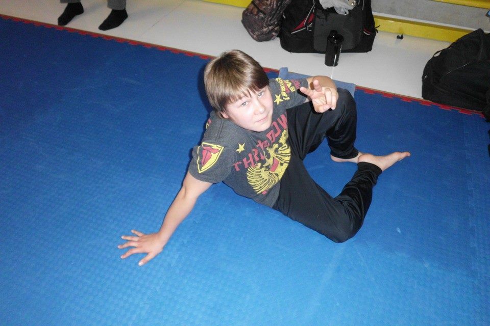 Ein Junge in Sportkleidung sitzt auf einer blauen Matte und macht ein Peace-Zeichen mit der rechten Hand.