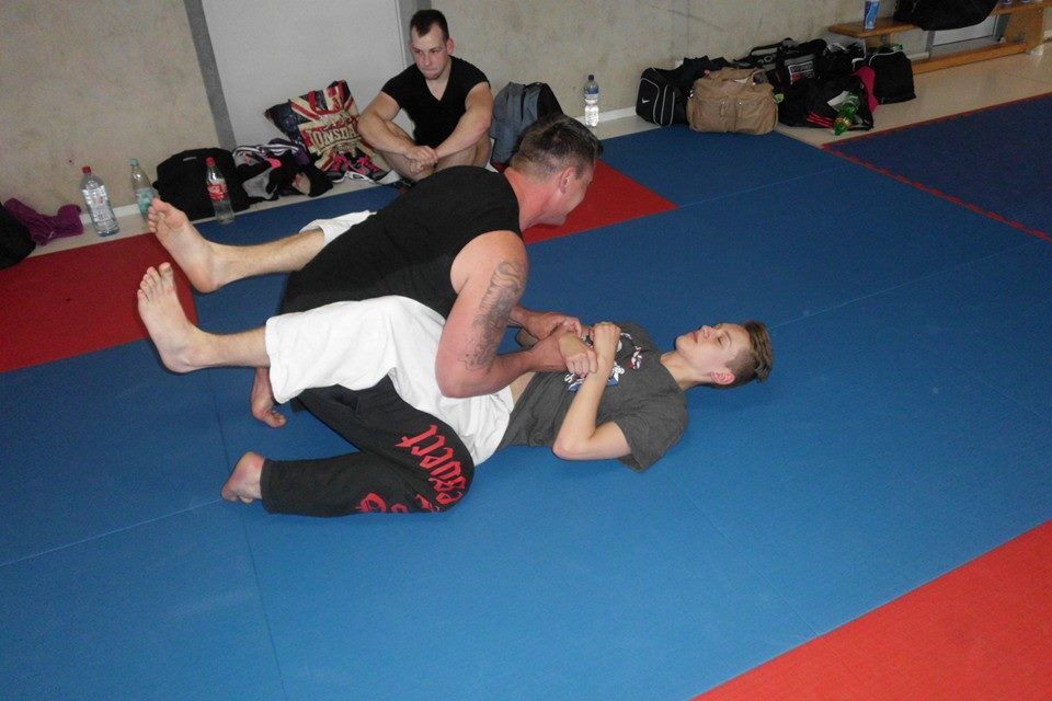 Zwei Personen &uuml;ben Bodenkampf auf einer blauen Trainingsmatte; Zuschauer im Hintergrund.