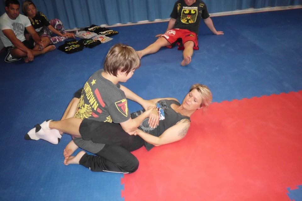 Zwei Personen trainieren auf einer Matte, w&auml;hrend Zuschauer im Hintergrund zuschauen. Eine Person sitzt auf der anderen.