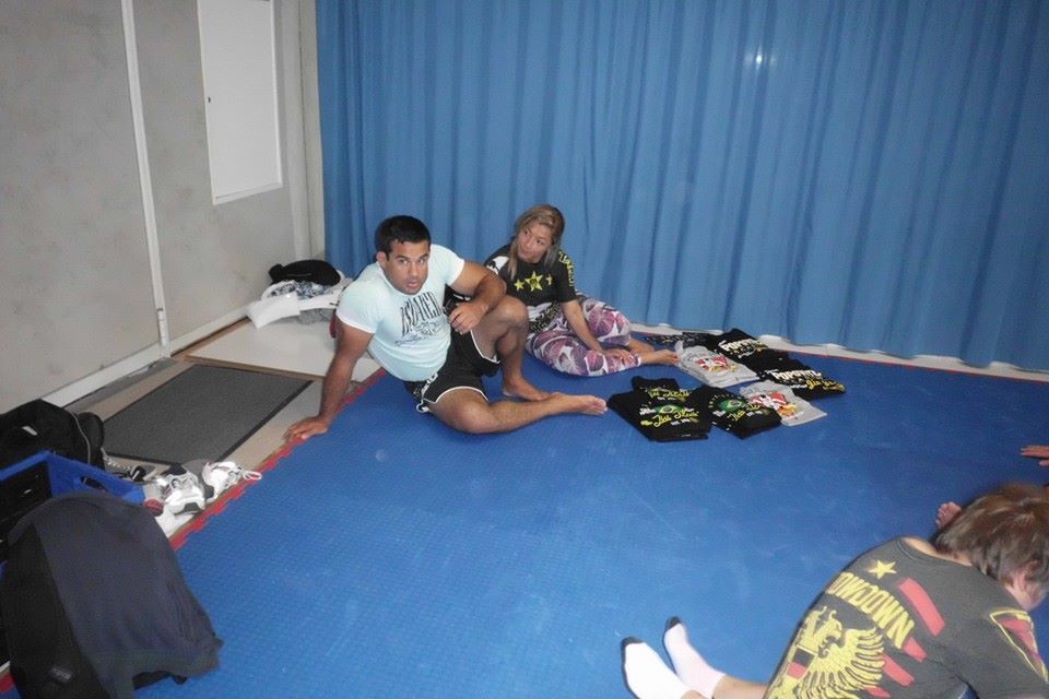 Zwei Personen sitzen auf einer blauen Mattenfl&auml;che, w&auml;hrend T-Shirts in der N&auml;he ausgebreitet sind.