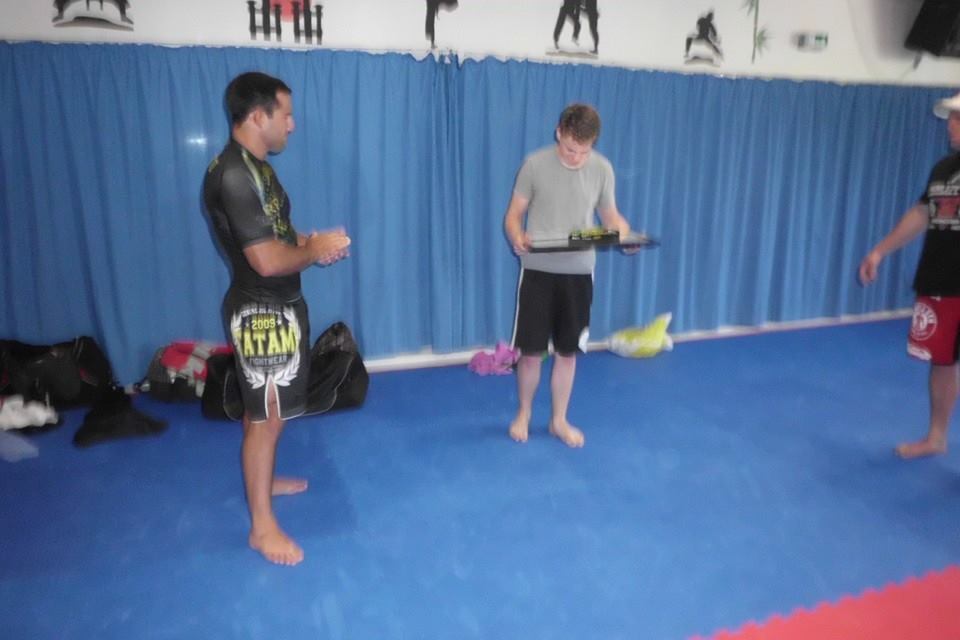 Drei Personen in Sportkleidung stehen auf einer blauen Matte in einem Trainingsraum mit blauer Vorhangwand.