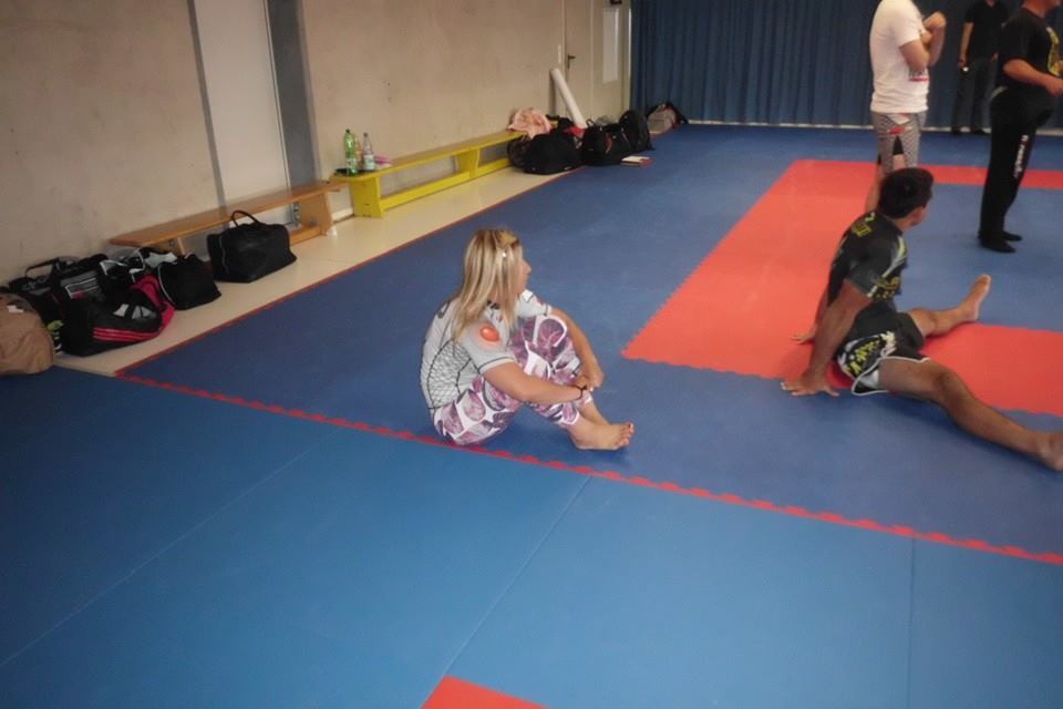 Eine Person sitzt auf einer blauen Matte in einem Trainingsraum, w&auml;hrend andere im Hintergrund &Uuml;bungen machen.