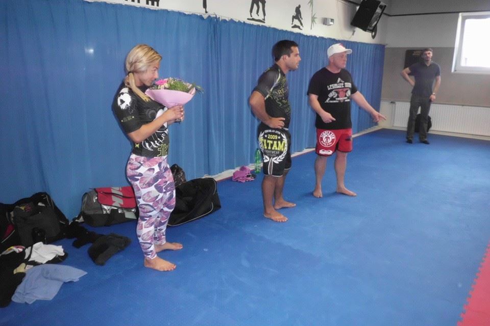 Vier Personen in Sportkleidung stehen in einem Trainingsraum mit blauer Matte und brauner Wandverkleidung.