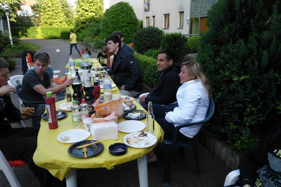 Gruppe von Personen an einem gedeckten Tisch im Freien, mit Essen und Getr&auml;nken, umgeben von gr&uuml;ner Vegetation.