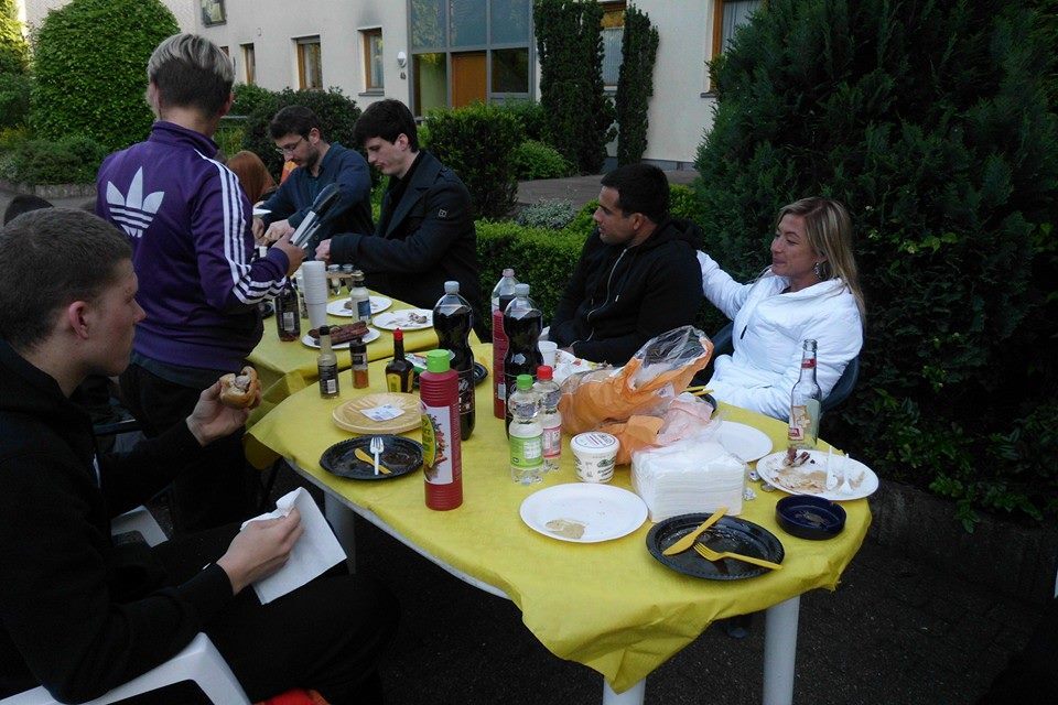 Gruppe von Personen sitzt an einem Tisch im Freien, umgeben von Essen und Getr&auml;nken auf einer gelben Tischdecke.