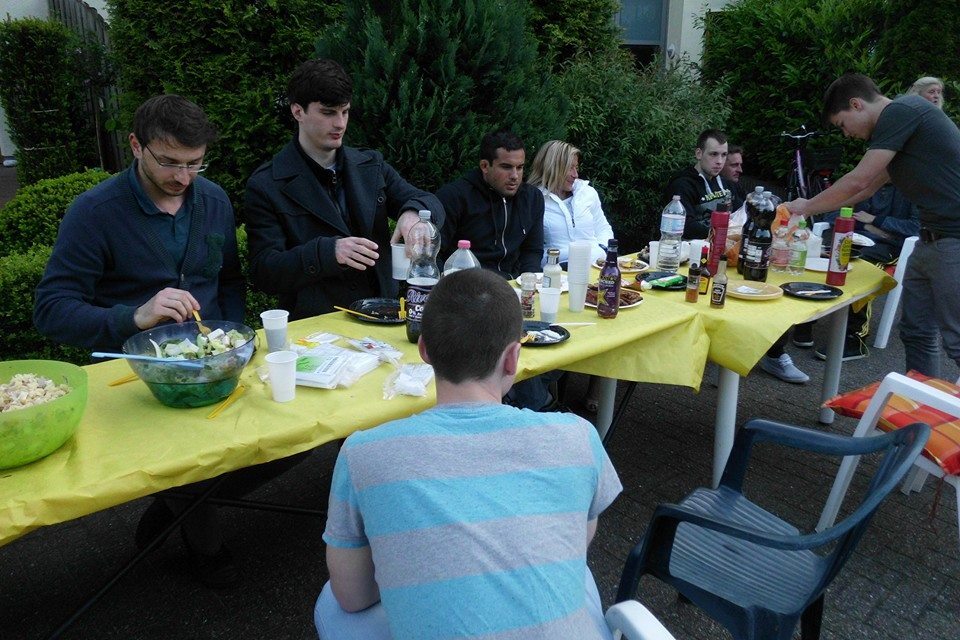 Sechs Personen sitzen an einem langen Tisch, der mit gelben T&uuml;chern, Tellern und Getr&auml;nken gedeckt ist.