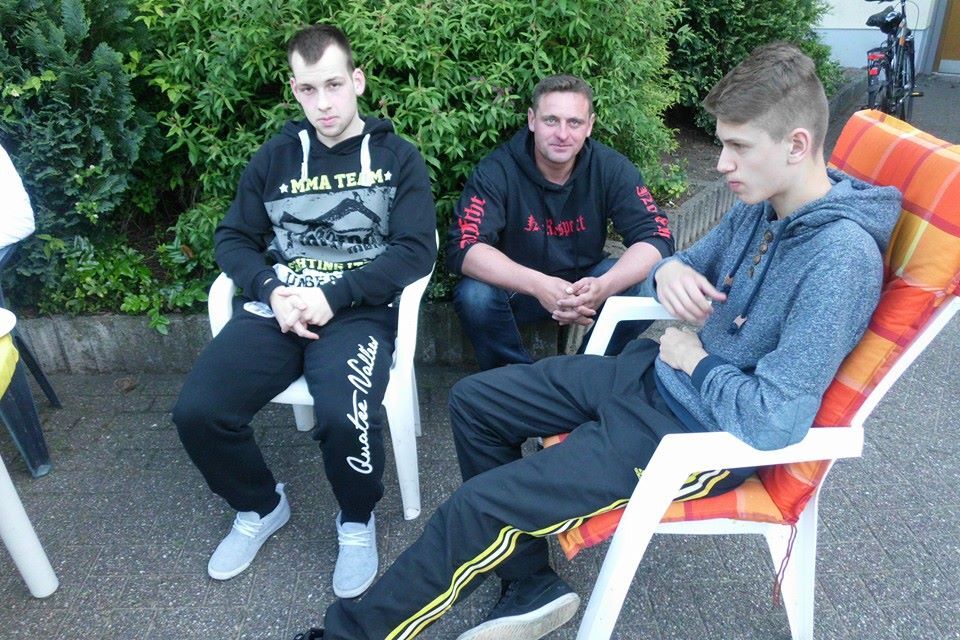 Drei junge M&auml;nner sitzen auf St&uuml;hlen im Freien, umgeben von Pflanzen und einer Terrasse.