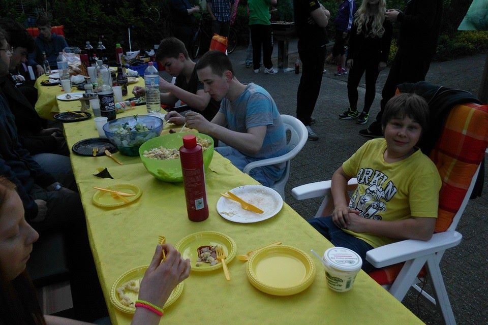 Tisch mit mehreren Personen, die essen und trinken; im Vordergrund junge Leute mit Tellern und Flaschen.