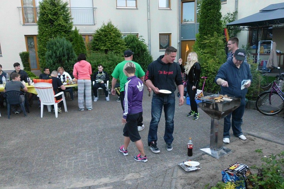 Grillparty im Freien mit mehreren Personen, die am Tisch sitzen und grillen, umgeben von B&auml;umen und Geb&auml;uden.