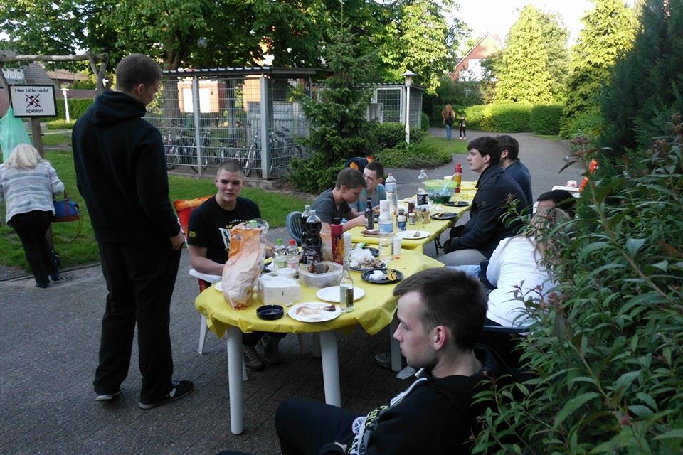 Gruppe junger Menschen sitzt an einem Tisch im Freien, umgeben von Essen und Getr&auml;nken, in einem Garten.