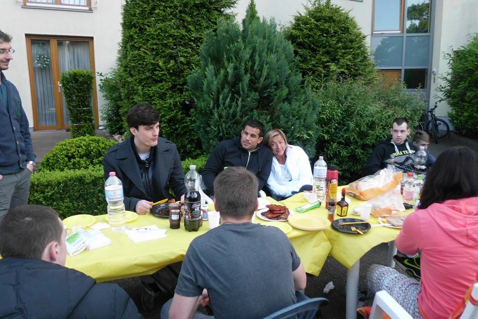 Gruppe von Personen sitzt an einem Tisch im Freien, umgeben von gr&uuml;nen Pflanzen, bei einem gemeinsamen Essen.