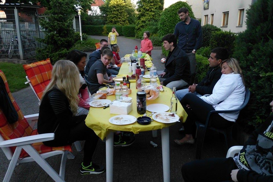 Gruppe von Personen bei einem Abendessen an einem langen Tisch im Freien, umgeben von B&auml;umen und Str&auml;uchern.