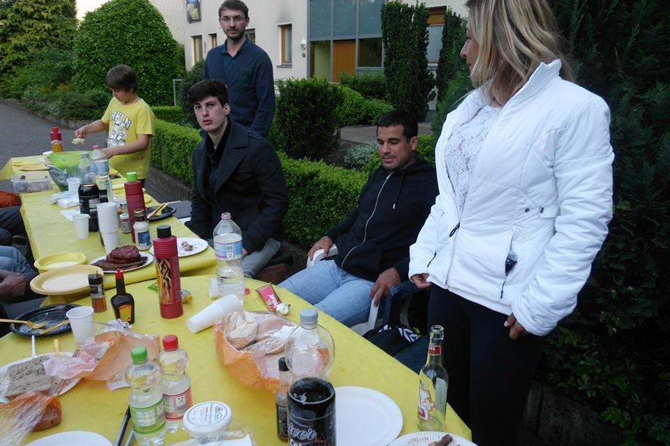 Verschiedene Personen sitzen und stehen an einem gedeckten Tisch im Freien, umgeben von Pflanzen und Getr&auml;nken.