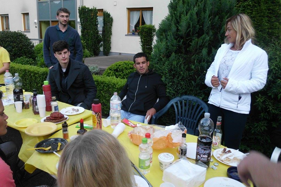 Gesellige Runde am Tisch mit Snacks und Getr&auml;nken, umgeben von gr&uuml;nen Str&auml;uchern, einige Personen unterhalten sich.
