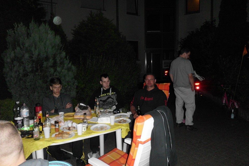 Gruppe von M&auml;nnern sitzt an einem Tisch im Freien, umgeben von Essen und Getr&auml;nken in der D&auml;mmerung.