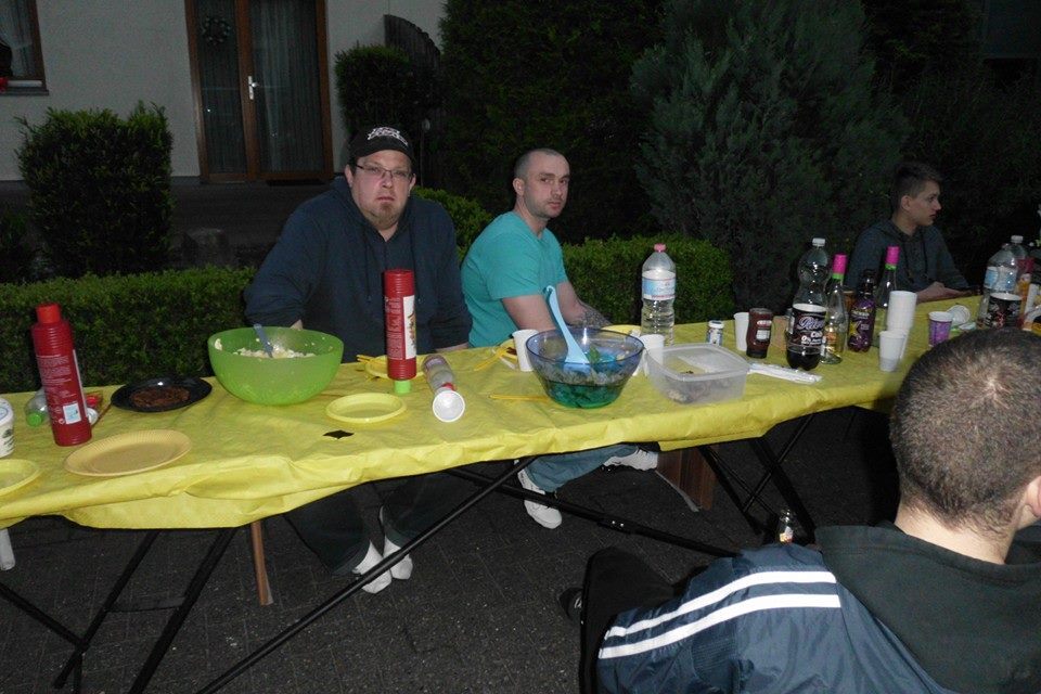 Gruppe von M&auml;nnern an einem gelben Tisch, umgeben von Snacks und Getr&auml;nken, im Freien bei Abendd&auml;mmerung.