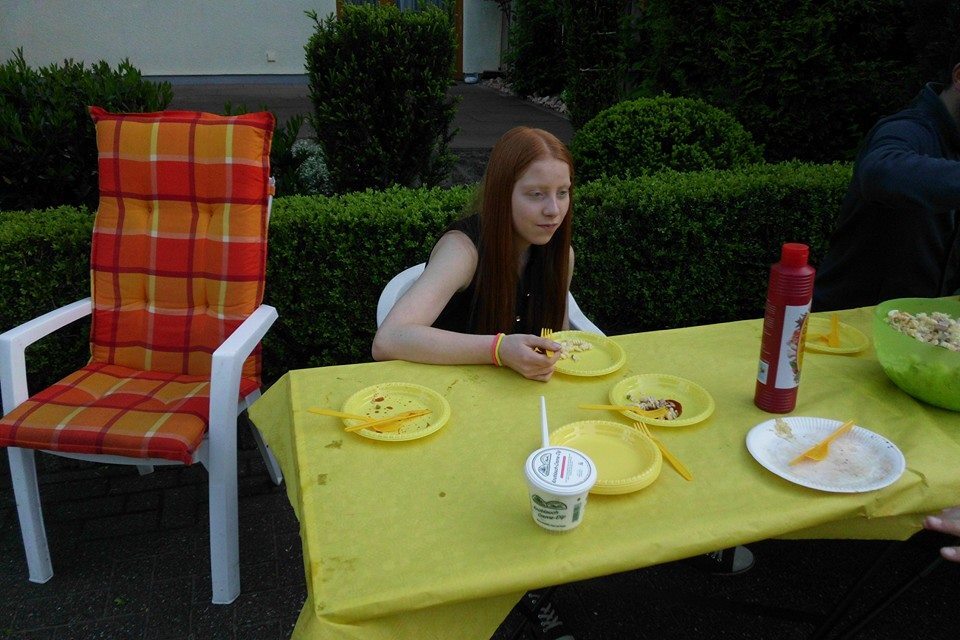 Junge mit rotem Haar sitzt an einem Tisch mit gelber Decke, umgeben von Essensgeschirr und einem Stuhl im Hintergrund.
