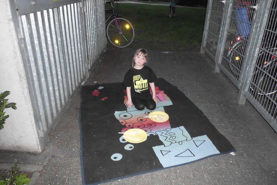 M&auml;dchen kniet auf einem bunten Teppich mit zwei Tellern Essen in einem begr&uuml;nten Au&szlig;enbereich. Fahrr&auml;der im Hintergrund.