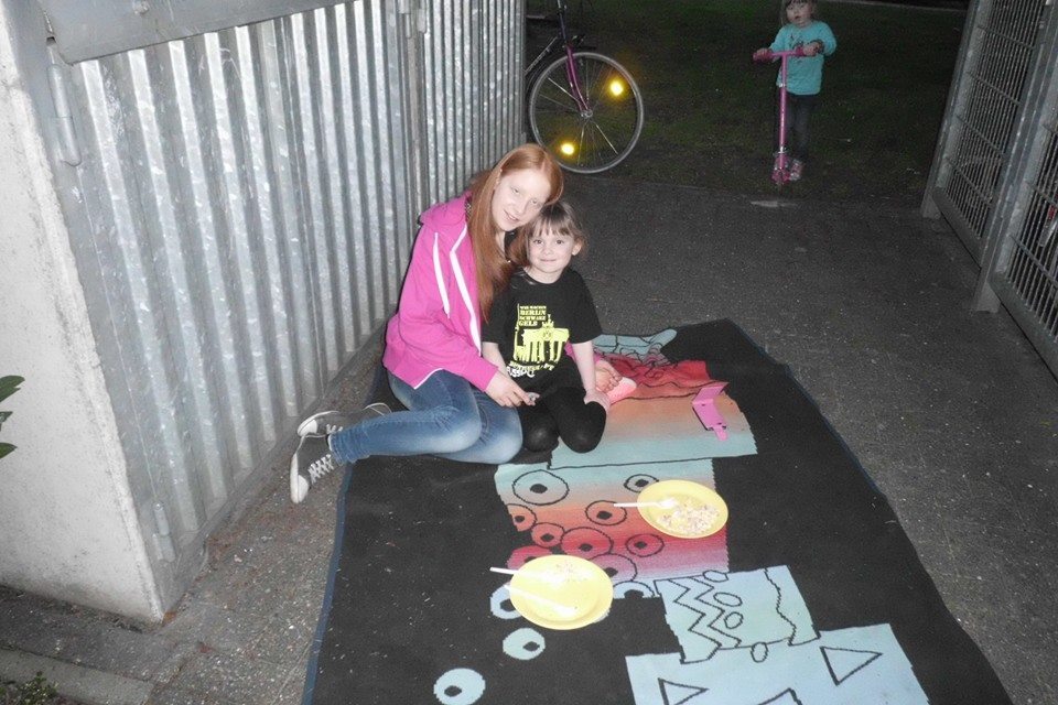Zwei Kinder sitzen auf einem bunten Spielteppich, w&auml;hrend eine weitere Person im Hintergrund steht. Teller mit Snacks sind sichtbar.