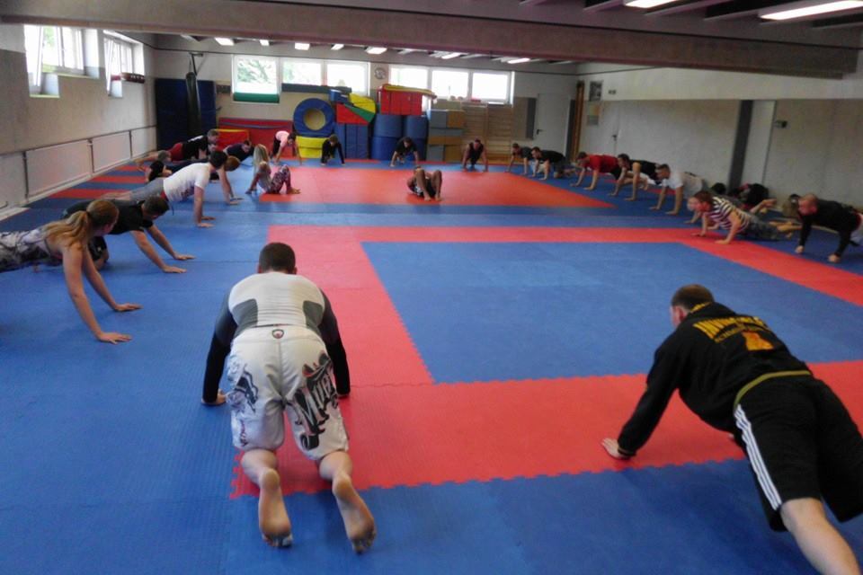 Trainingseinheit in einer Sporthalle mit mehreren Personen, die auf einer blauen Matte Liegest&uuml;tze machen.