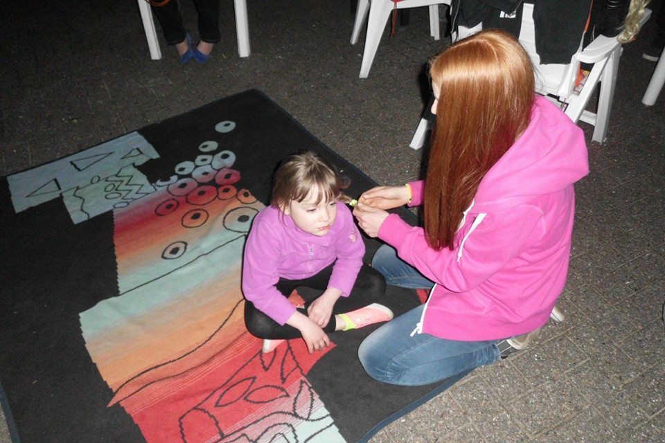 Ein M&auml;dchen sitzt auf einem bunten Teppich, w&auml;hrend eine Frau ihr die Haare flechtet.
