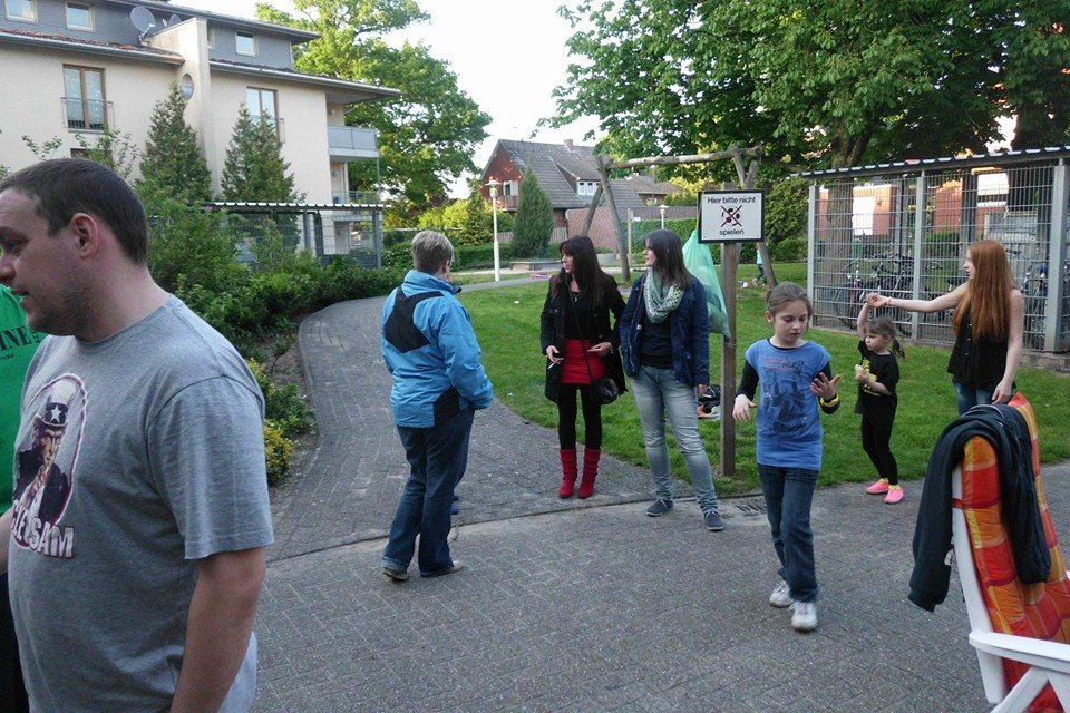 Gruppe von f&uuml;nf Personen auf einem Weg in einem Garten, umgeben von B&auml;umen und Wohngeb&auml;uden, mit Spielverbotsschild.