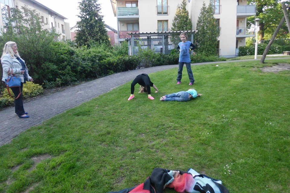 Kinder spielen auf einer Wiese: ein Kind macht einen Handstand, andere liegen oder stehen in der N&auml;he. Eine Frau beobachtet.