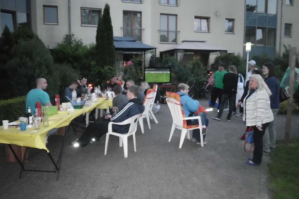 Personen sitzen an einem Tisch im Freien, umgeben von Pflanzen, w&auml;hrend ein Fu&szlig;ballspiel auf einem Fernseher l&auml;uft.