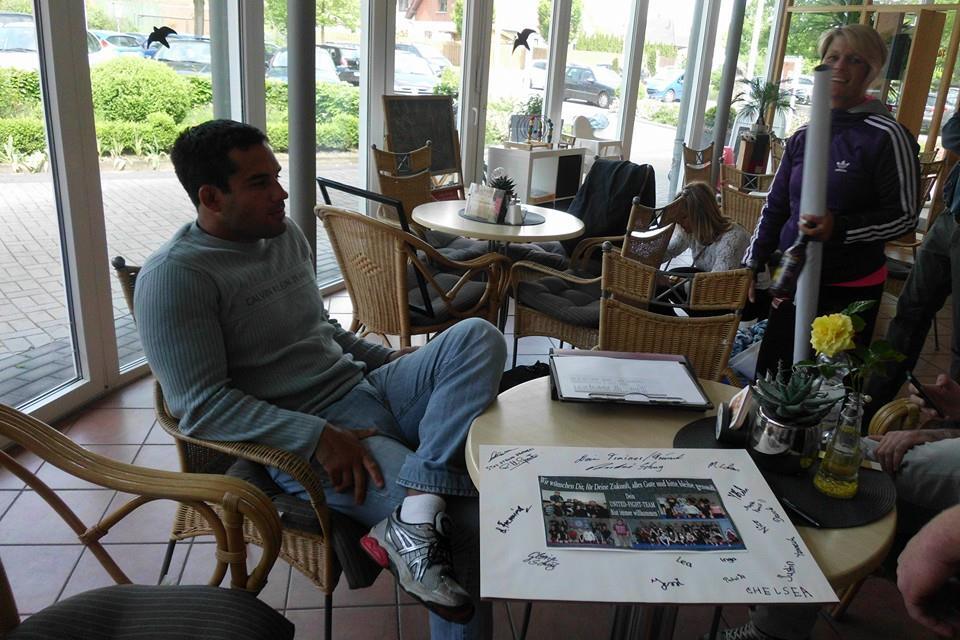 Mann sitzt entspannt in einem Caf&eacute;, umgeben von Personen und einem Tisch mit einem signierten Poster.