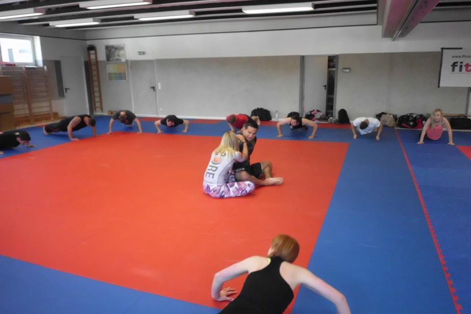 Gruppentraining auf einer blauen und roten Mattenfl&auml;che mit Teilnehmern in verschiedenen &Uuml;bungen und Paararbeit im Vordergrund.