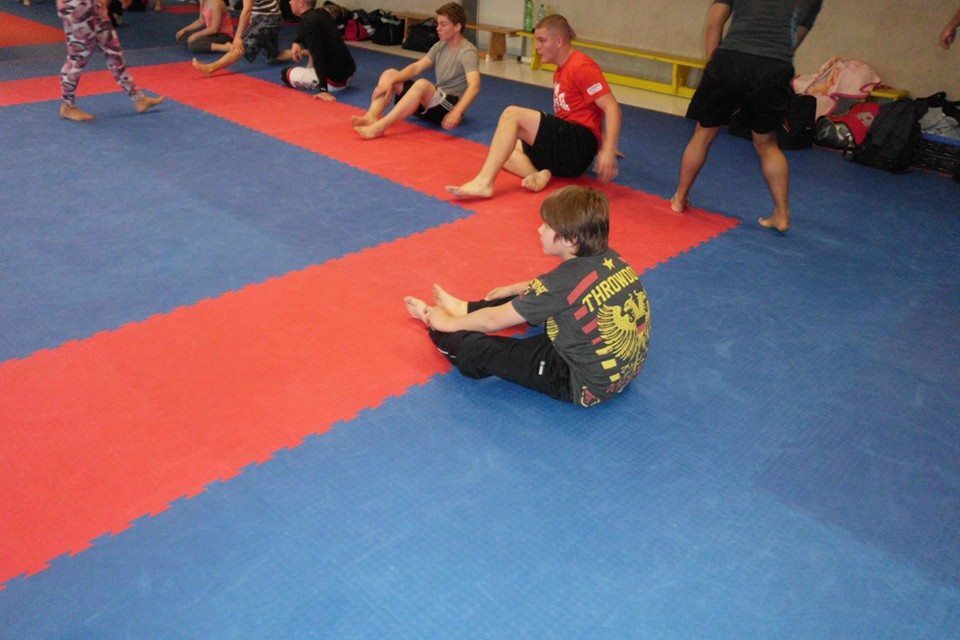 Gruppe von Jugendlichen auf einer Sportmatte beim Training, einige dehnen sich, andere sitzen in verschiedenen Positionen.