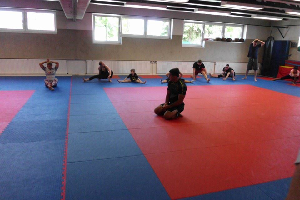 Gruppentraining auf einer blauen Matte, Teilnehmer dehnen und sitzen in einer Kampfsporthalle.