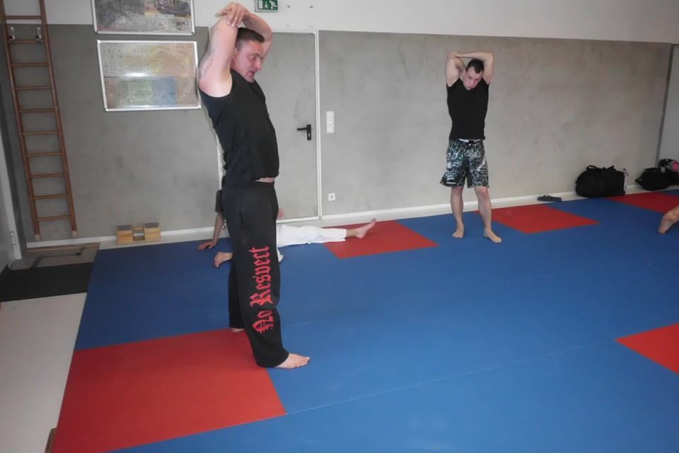 Zwei M&auml;nner dehnen in einer Kampfsporthalle auf blauen Matten, im Hintergrund liegt eine Person auf dem Boden.