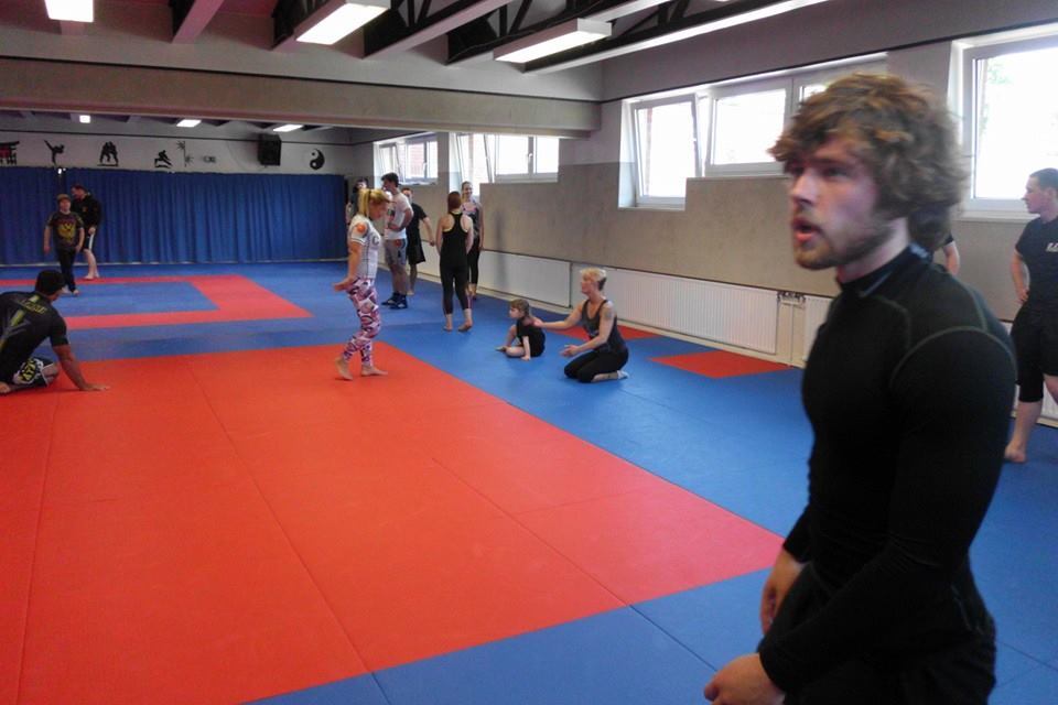 Trainingsszene in einem Kampfsportstudio mit Personen auf blauen und roten Matten. Einige trainieren, andere beobachten.