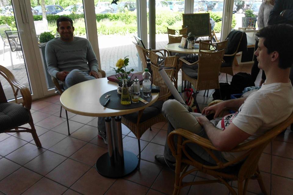 Zwei M&auml;nner sitzen in einem Caf&eacute;, der eine l&auml;chelt, der andere liest an einem Tisch mit Gl&auml;sern und einer Blume.