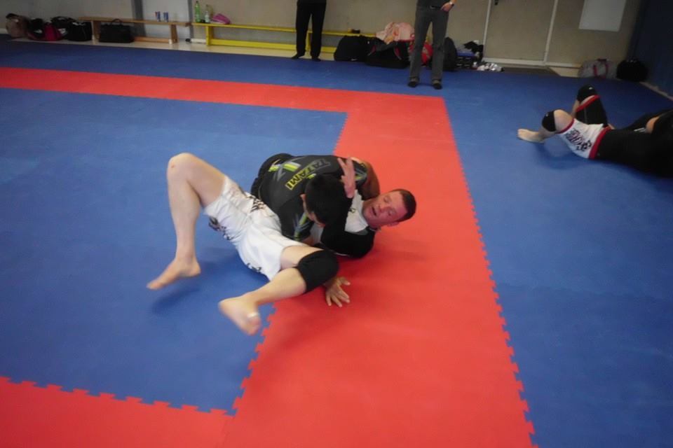 Zwei Sportler &uuml;ben auf einer blauen Matte; einer f&uuml;hrt eine Technik aus, w&auml;hrend der andere im Hintergrund liegt.