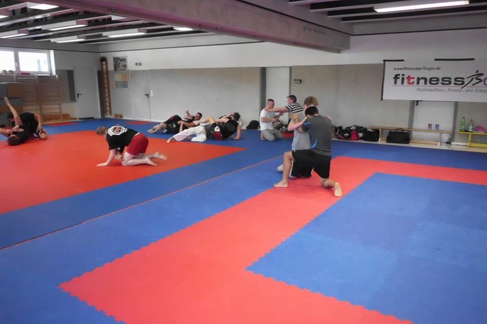 Trainingseinheit in einem Fitnessraum mit mehreren K&auml;mpfern auf blauen und roten Matten in verschiedenen Positions&uuml;bungen.