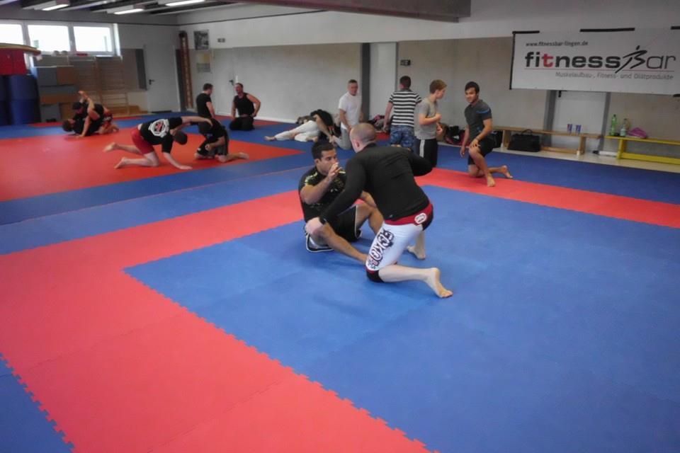 Trainingsszene in einem Kampfsportraum mit mehreren Personen auf einer blauen und roten Matte.