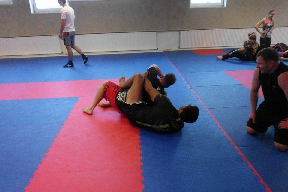 Zwei K&auml;mpfer im Bodenkampf auf blauen und roten Matten in einem Trainingsraum. Zuschauer im Hintergrund.