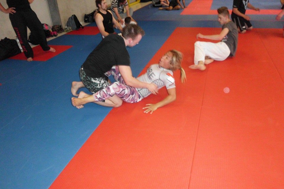 Zwei Sportler trainieren Bodentechniken auf einer Matte in einem Kampfsporttraining. Im Hintergrund sind weitere trainierende Personen.