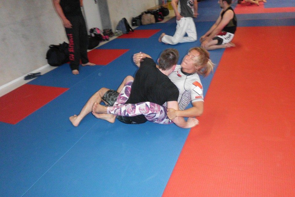Zwei Personen &uuml;ben Grappling-Techniken auf blauen und roten Matten in einem Kampfsporttraining.