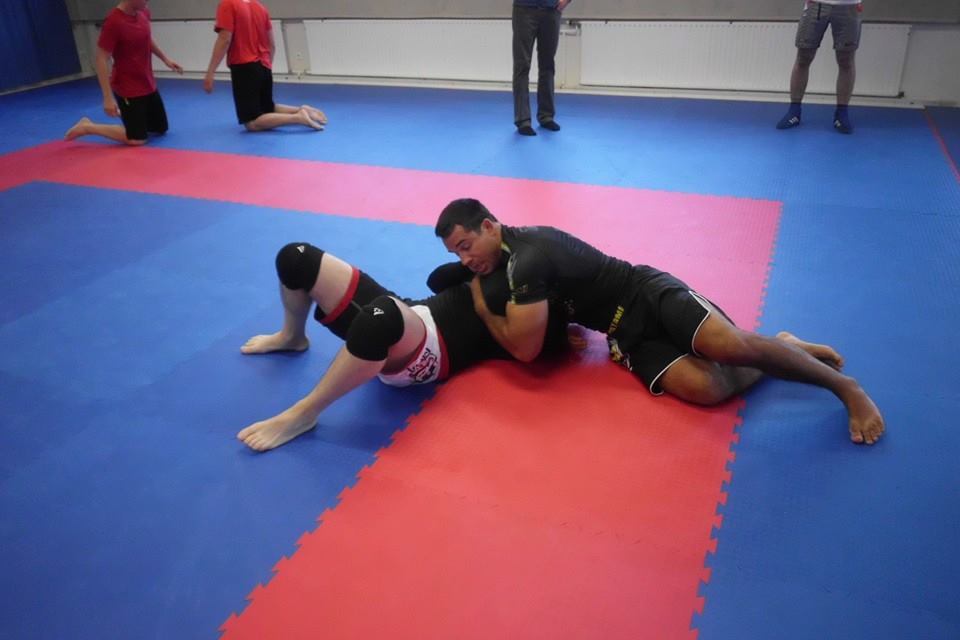 Zwei Kampfsportler &uuml;ben Bodentechniken auf einer blauen und roten Mattenfl&auml;che. Weitere Teilnehmer im Hintergrund.