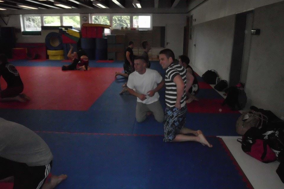 Sportliche Trainingsszene in einer Turnhalle mit mehreren Personen auf blauen und roten Matten, einige &uuml;ben k&auml;mpferische Techniken.