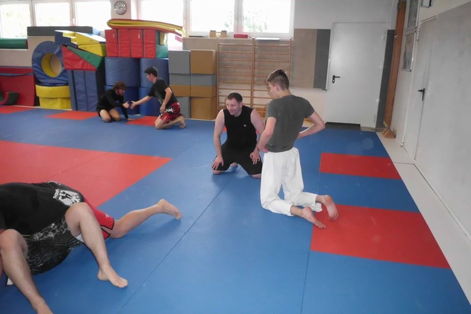 Leere Sporthalle mit mehreren Personen, die auf blauen Matten trainieren, w&auml;hrend andere in der N&auml;he entspannen.