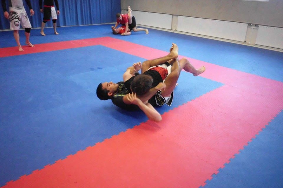 Zwei Sportler praktizieren Grappling auf einer blau-roten Matte in einem Trainingsraum.