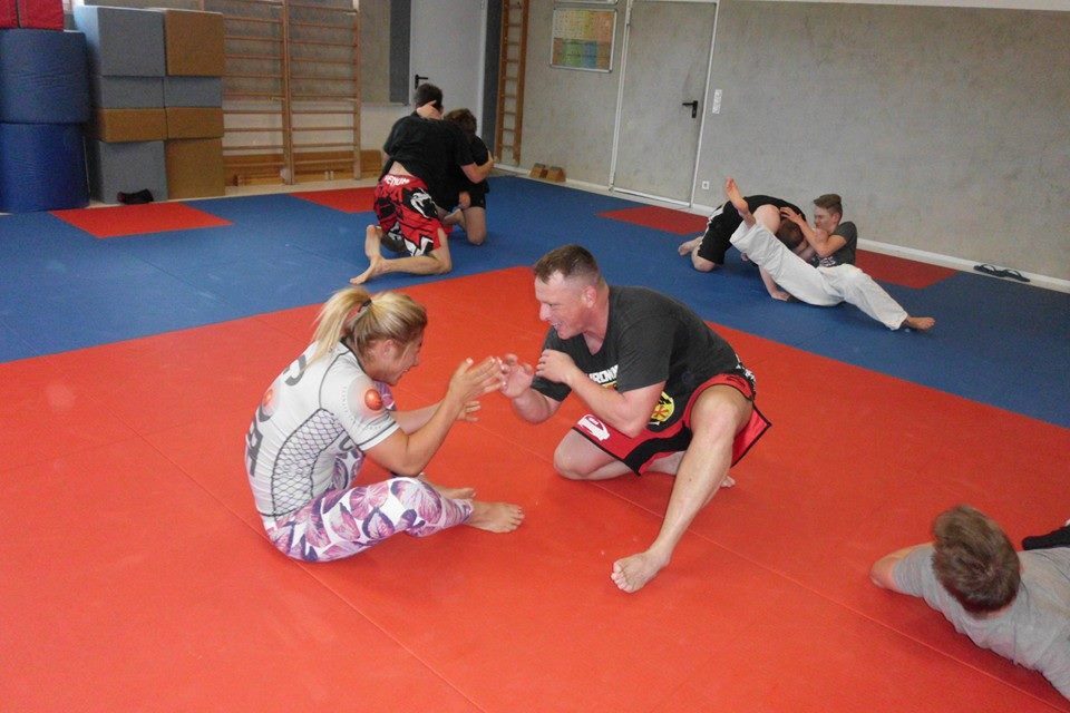 Zwei K&auml;mpfer &uuml;ben auf einer roten Matte, w&auml;hrend weitere Teilnehmer im Hintergrund verschiedene Techniken trainieren.