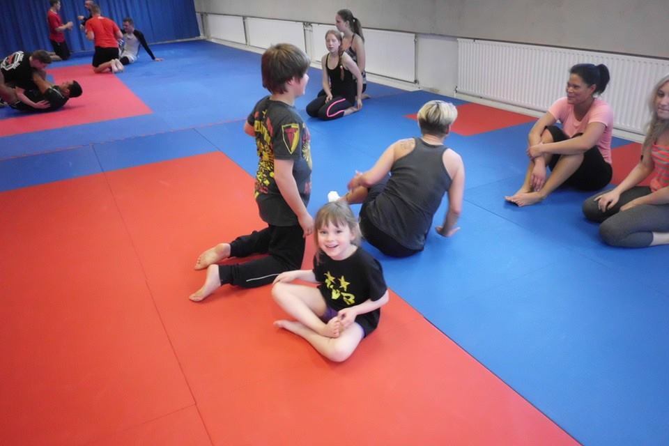 Kinder und Erwachsene &uuml;ben auf blauen und roten Matten; ein Junge sitzt l&auml;chelnd im Vordergrund.